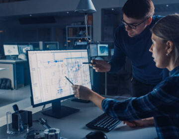 Engineer Working on Desktop Computer, Screen Showing CAD Software with Technical Blueprints, Her Male Project Manager Explains Job Specifics. Industrial Design Engineering Facility Office