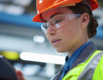 Portrait supervisor engineer working in safety uniform in manufacturing company. Focused brunette industrial engineer woman in safety glasses looking tablet computer. Online factory technology concept